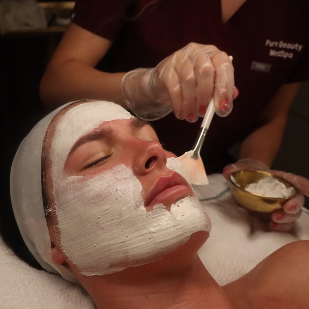 Woman receiving a facial with a thick white mask being applied to her face by an esthetician wearing gloves, lying spa bedside.