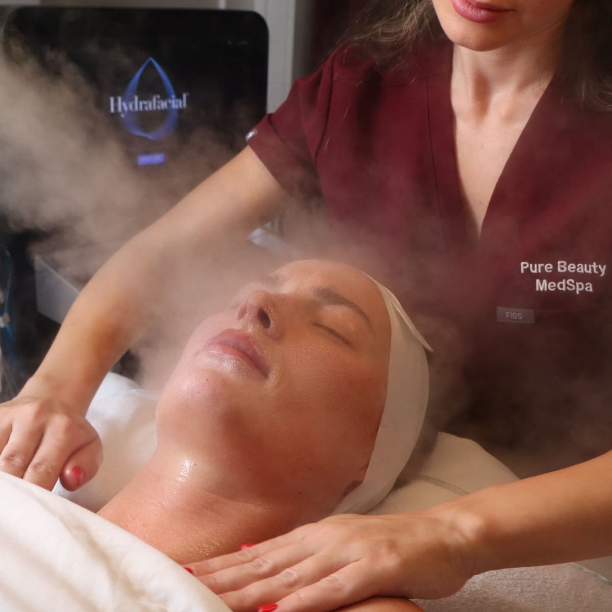 A woman receiving a steam facial in a spa, eyes closed while a practitioner uses steam over her face.