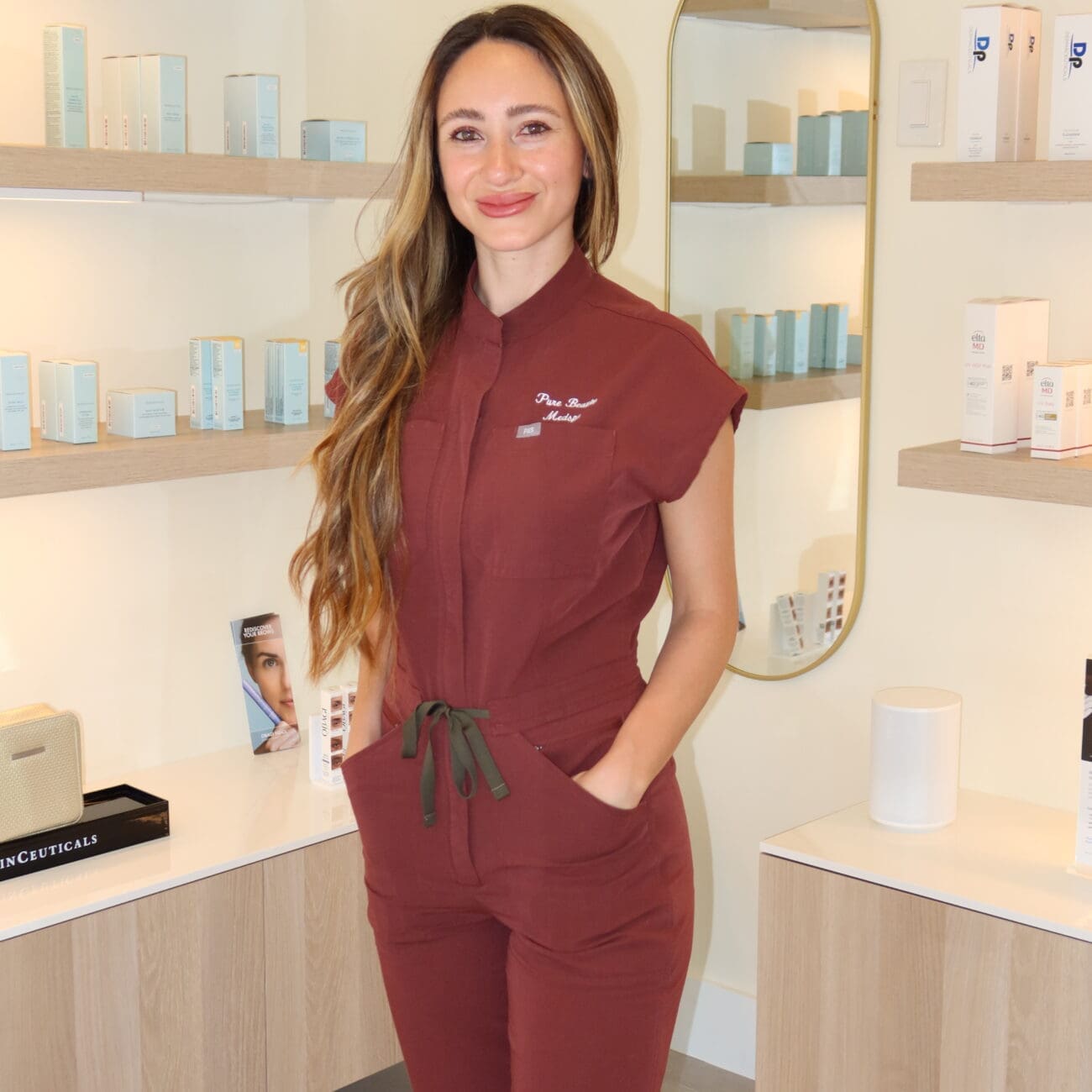 Smiling woman in burgundy scrubs stands in a clean skincare shop with shelves of light blue boxes behind her.